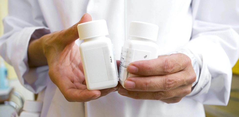 Foto em detalhe das mãos de um profissional na frente do seu corpo. A pessoa segura dois recipientes de produto de plástico branco com tampa. 